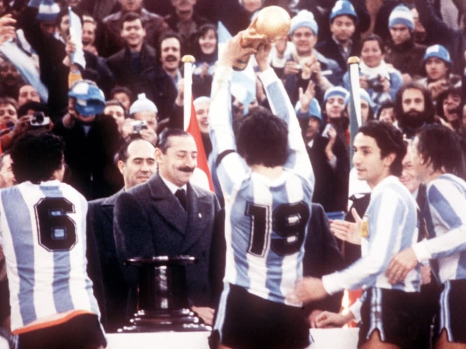 Mit dem Rücken zur Kamera: Vier argentinische Spieler. Im Hintergrund Videla, der mit Kittel und Krawatte etwas lächelt.