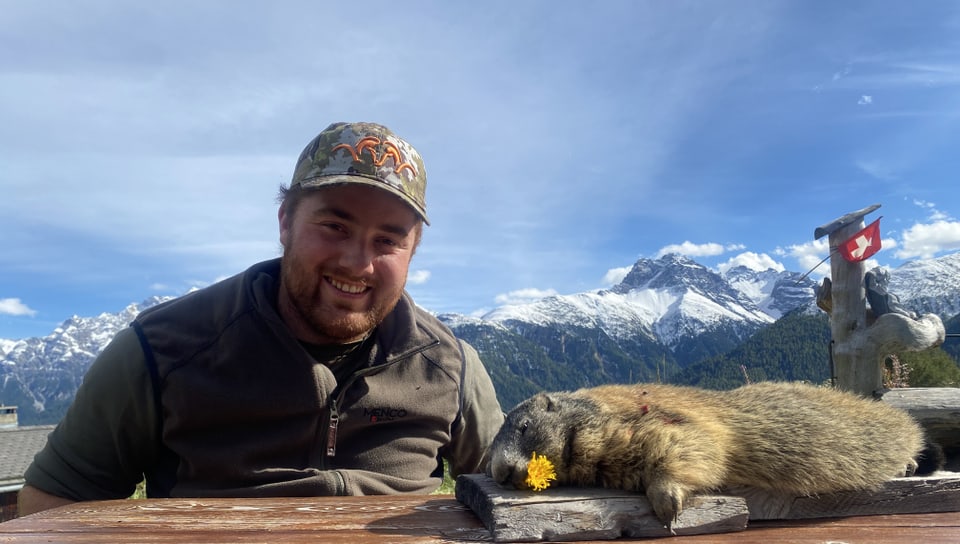 Lächelnder Mann neben einem erlegten Murmeltier vor Bergkulisse.