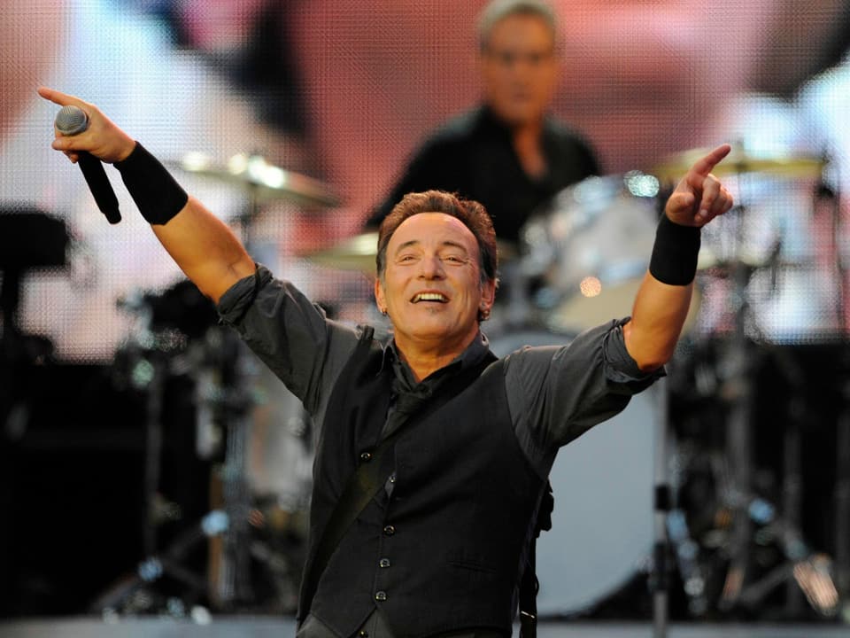 Bruce Springsteen steht lachend während einem Konzert auf einer Bühne und zeigt mit beiden Händen ins Publikum.