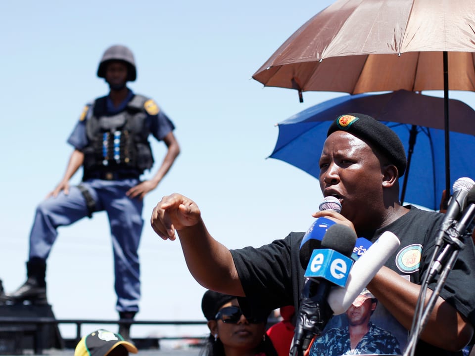 Julius Malema hält eine Rede vor Minenarbeitern. Im Hintergrund ein Polizist.
