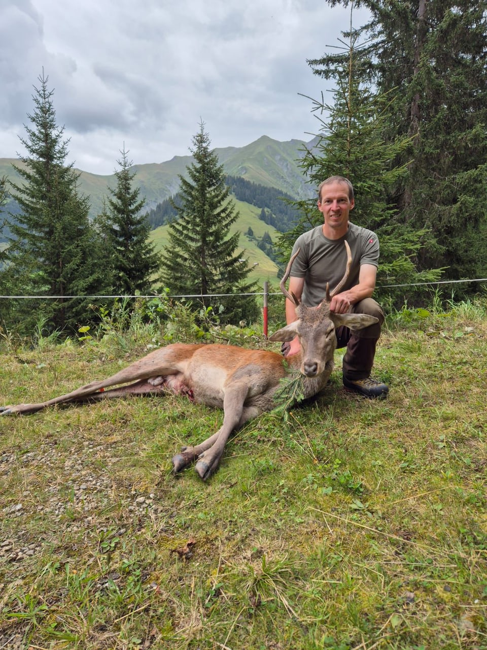 Mann posiert neben erlegtem Hirsch im Wald mit Bergen im Hintergrund.