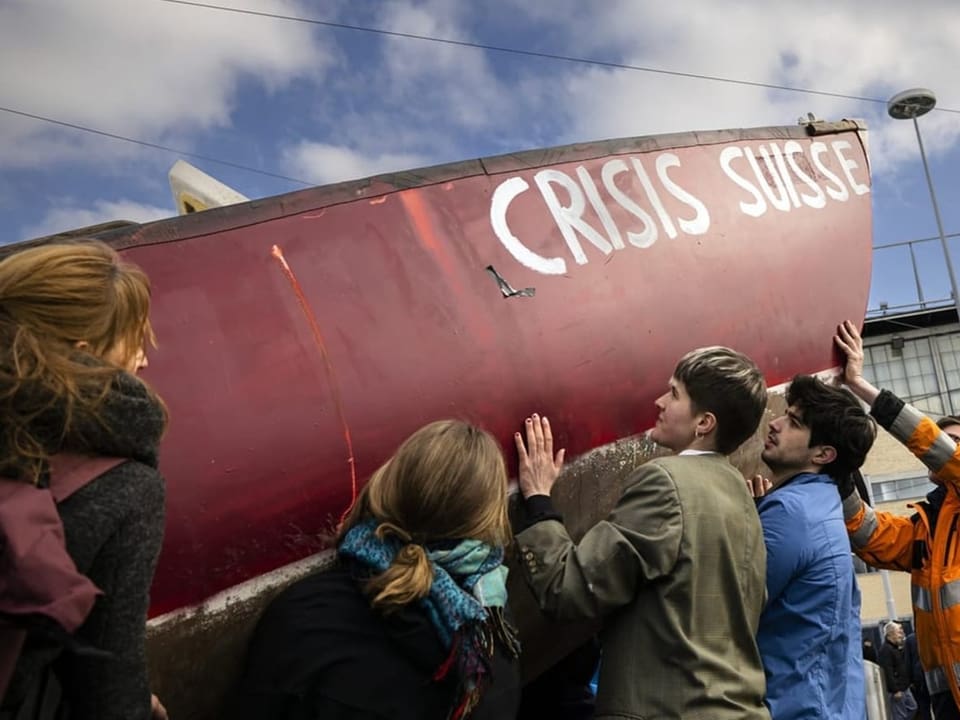 Mehrere Personen heben einen Bootsrumpf mit der Aufschrift «Crisis Suisse» an.