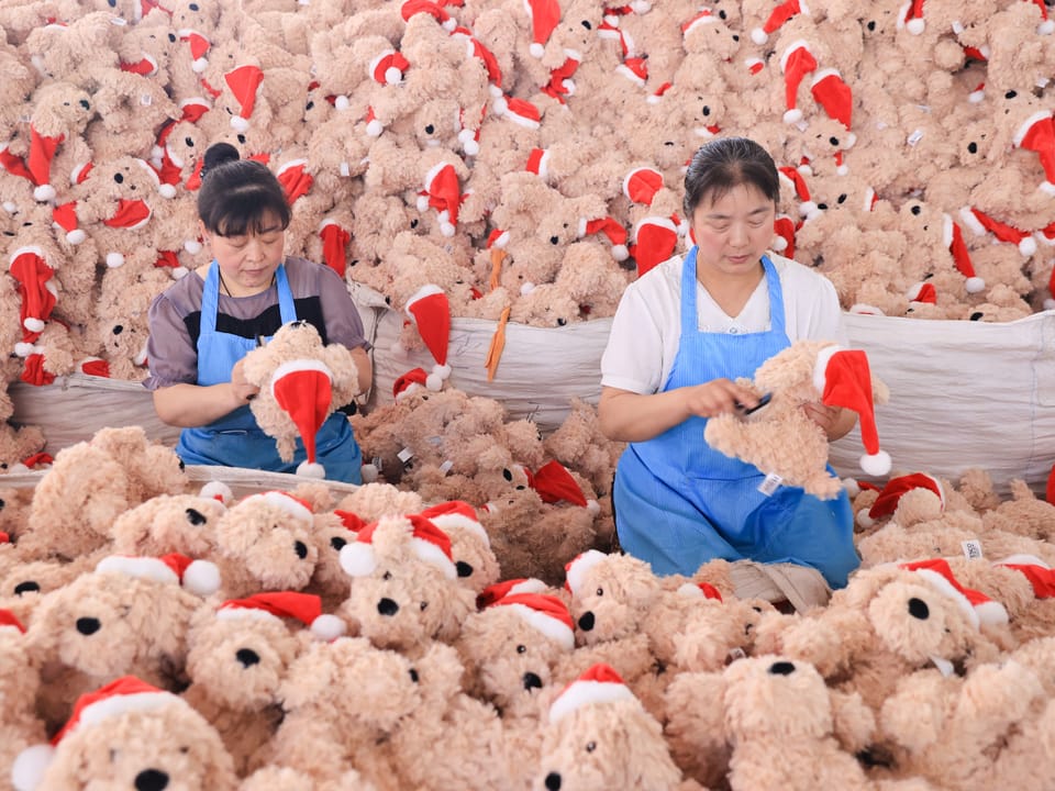 Zwei Arbeiterinnen montieren Weihnachtsmützen an Plüschbären in einer Fabrik.