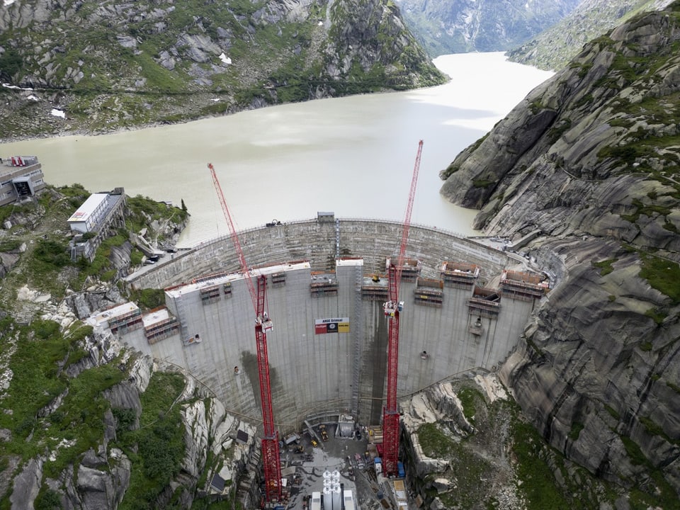 Luftaufnahme eines Staudamms mit Baukränen in einer bergigen Landschaft.