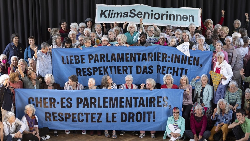 Gruppe älterer Frauen hält Banner mit Aufschriften und 'Liebe Parlamentarier:innen, respektiert das Recht!'