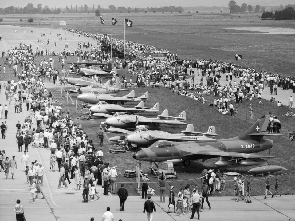 Kampfflugzeuge stehen nebeneinander auf dem Flugplatz. Flugfans stehen herum und bestaunen die Flugzeuge. 