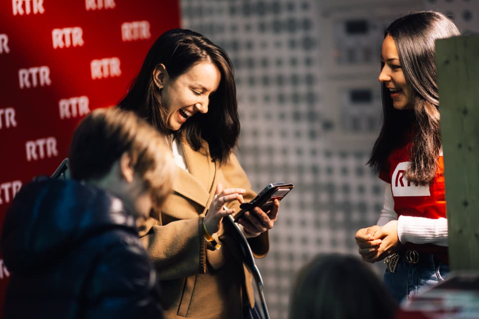 Zwei Frauen lachen und schauen auf ein Smartphone, drinnen vor rotem Hintergrund.