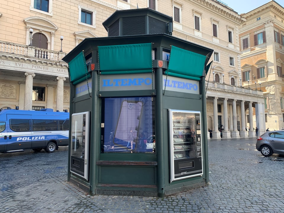 Grüner Zeitungskiosk wie Getränkeautomat auf einem Platz in Rom mit Polizeifahrzeug im Hintergrund.