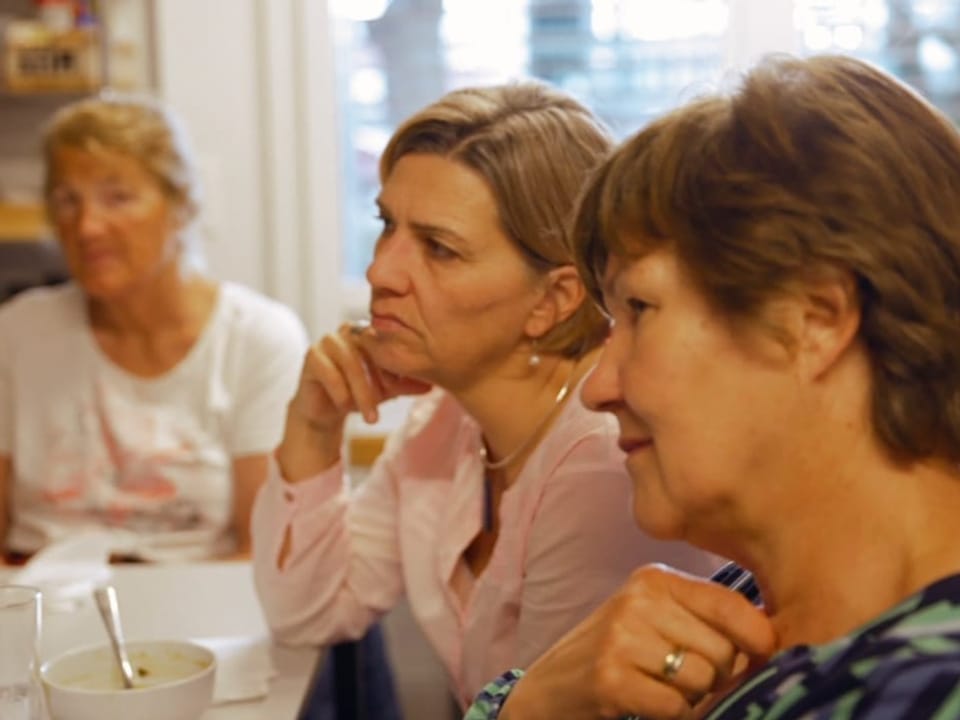 Drei Frauen sitzen an einem Tisch und hören einer Person zu, die nicht im Bild zu sehen ist.