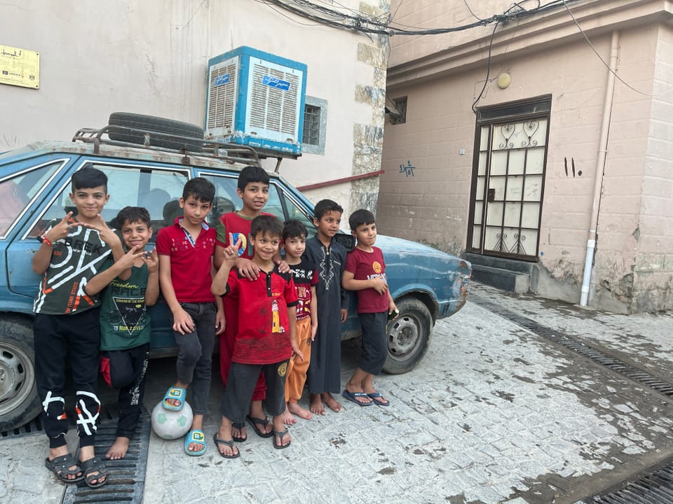 Gruppe von Kindern steht vor einem alten Auto auf einer gepflasterten Strasse.
