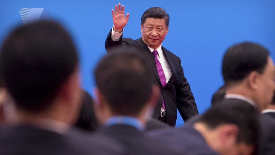 Der Präsident Xi grüsst mit der Hand und steht über einer verschwommenen Menschenmenge.
