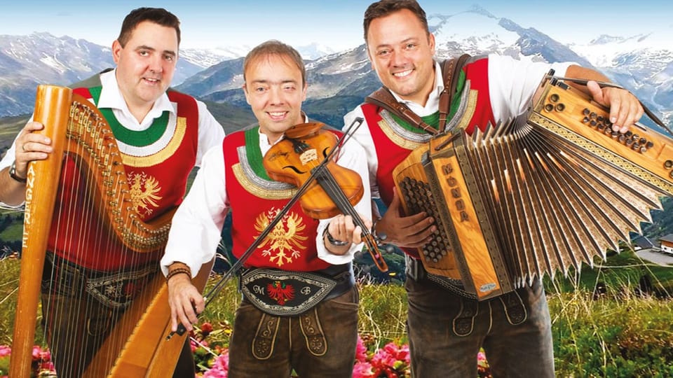 Ursprung Buam in Tiroler Trachten mit Instrumenten vor Bergkulisse.