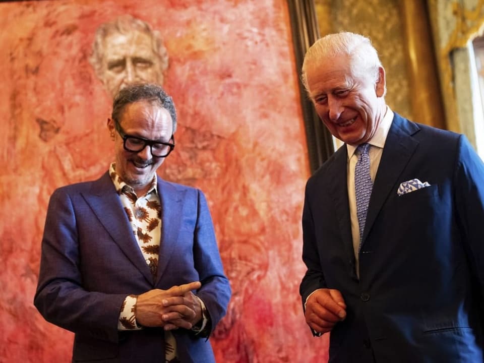 Zwei Männer in Anzügen vor einem grossen roten Gemälde, lächelnd.
