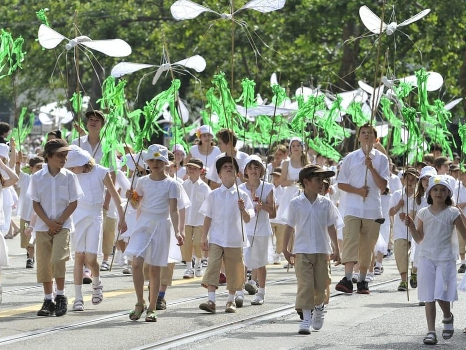 Eine grosse Gruppe von Kindern mit weissem Oberteil und beigen Hosen. Sie halten Stäbe mit grünen Lamellen in die Höhe. 