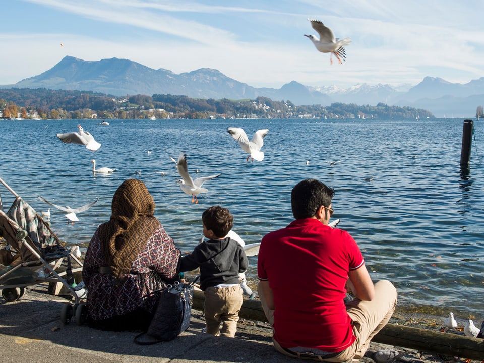 Eine Frau mit Kopftuch, ein Kind und ein Mann sitzen am Seeufer in Luzern.