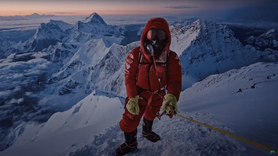 Inoxtag mit Sauerstoffmaske auf dem Berg. Hinter ihm schneebedeckte Gipfel.