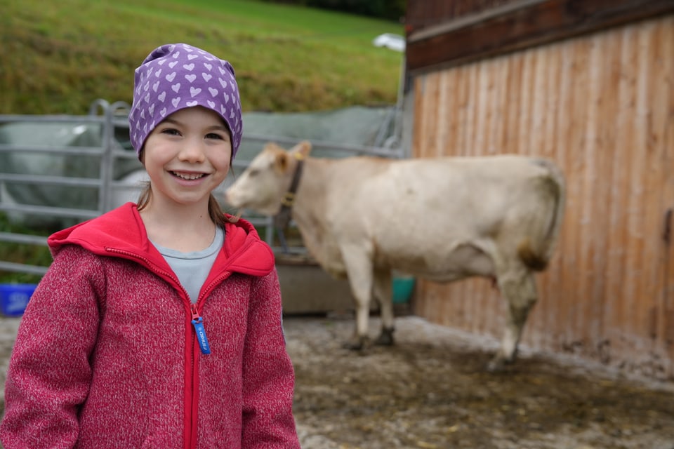 Lächelndes Mädchen in lila Mütze und rotem Pullover steht vor einer Kuh.