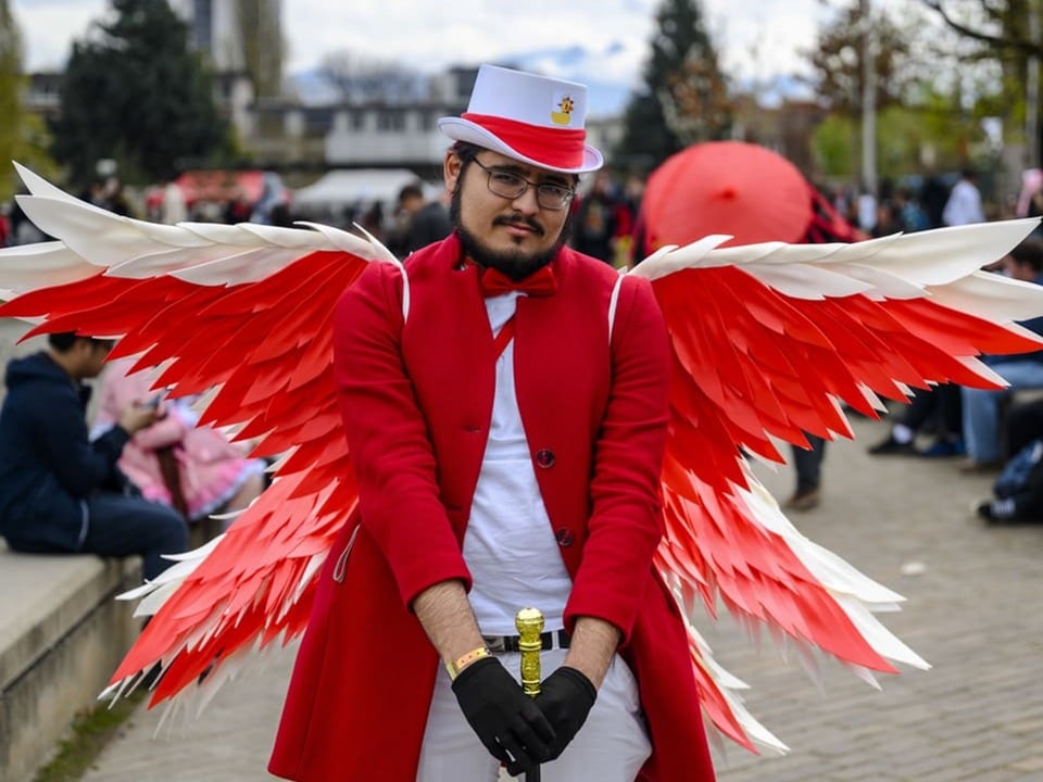 Mann in rotem Mantel und weissen Hut trägt rote und weisse Flügel am Rücken und schaut frontal in die Kamera.
