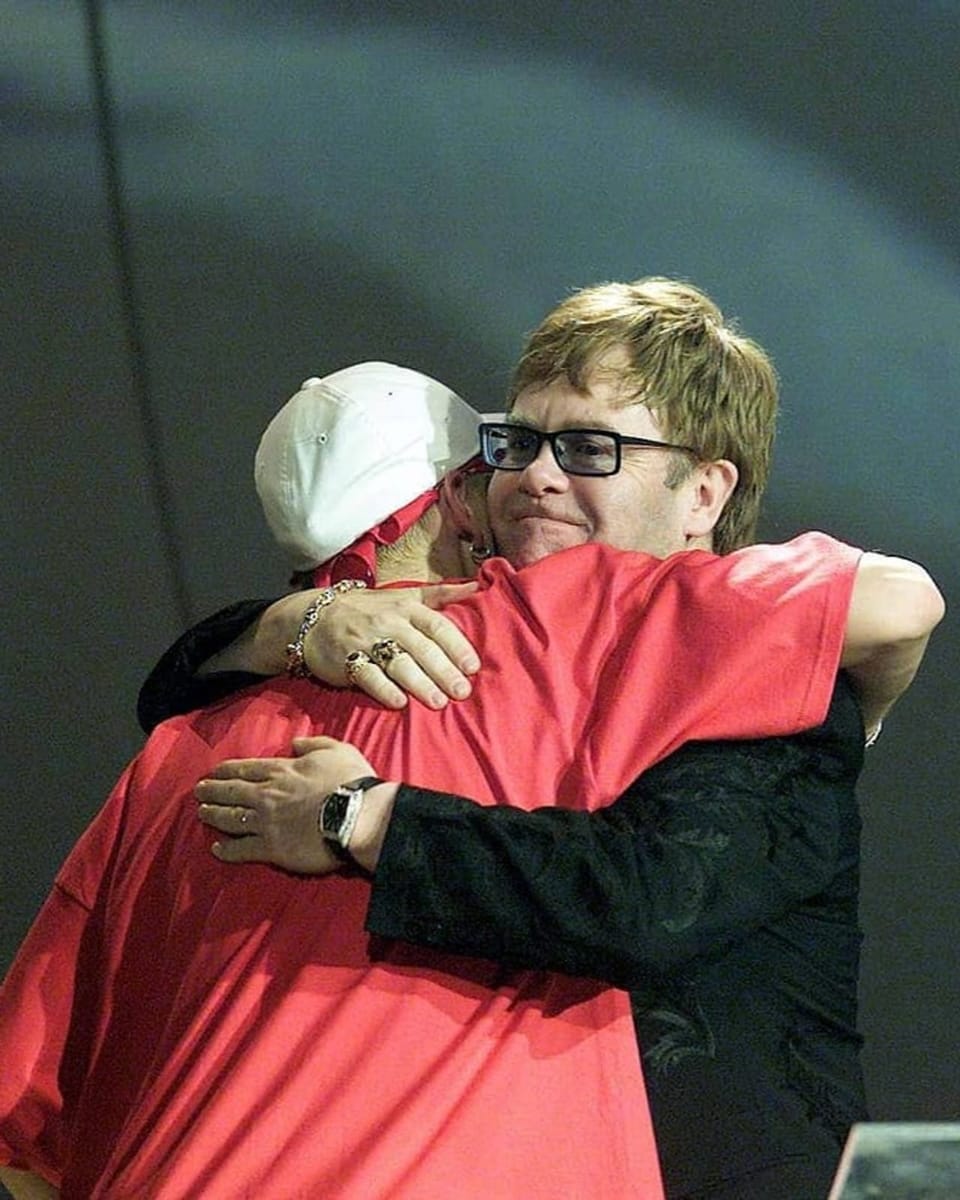 Eminem und Elton John umarmen sich. Eminem ist mit dem Rücken zu sehen und trägt ein rotes T-Shirt.