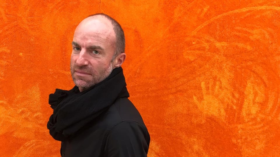 Ein Mann mittleren Alters mit schwarzem Pulli und schwarzem Schal steht vor einer orangen Wand.