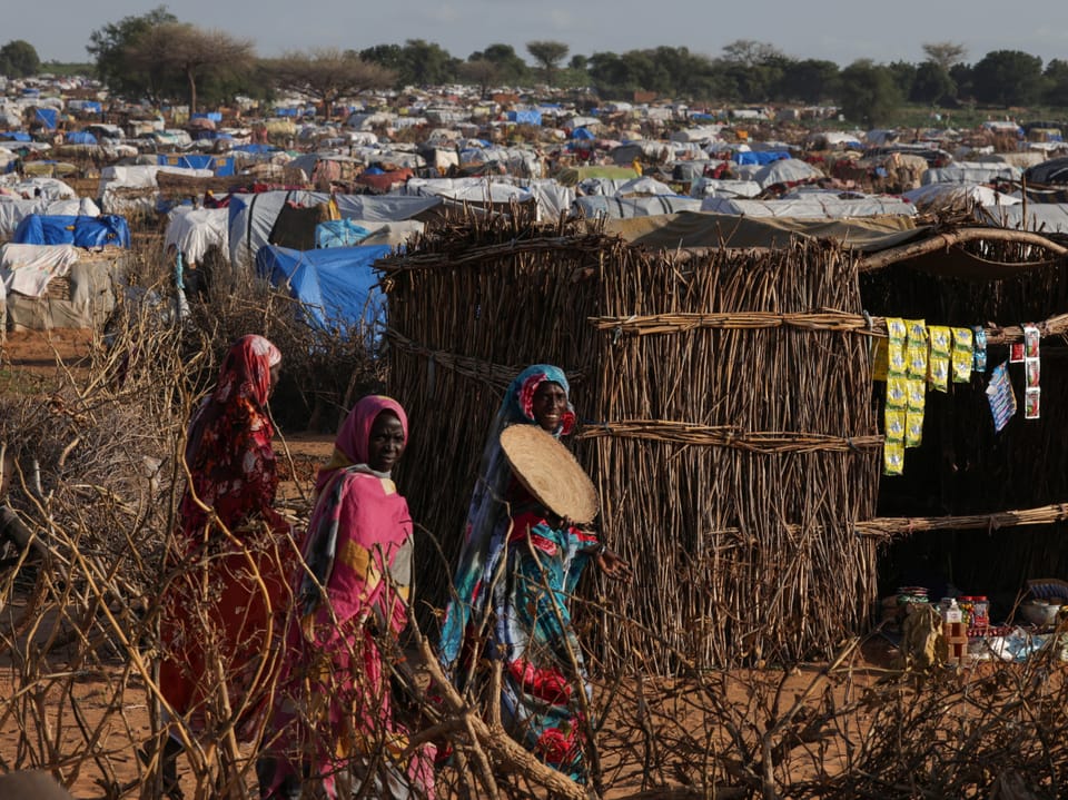 Frauen in bunten Kleidern gehen an Strohhütte in einem Flüchtlingslager vorbei.