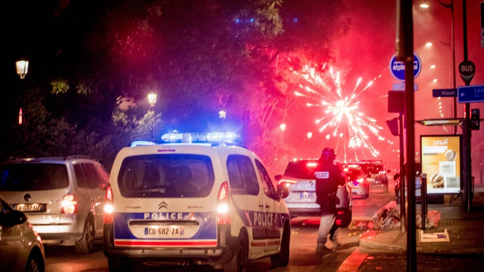 Ein Polizeiauto. Dahinter ein rotes Feuerwerk.