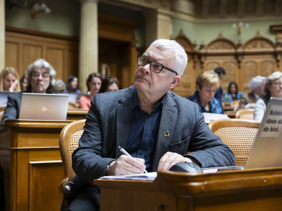 Ein älterer Mann sitzt in einem Sitzungssaal und macht Notizen.