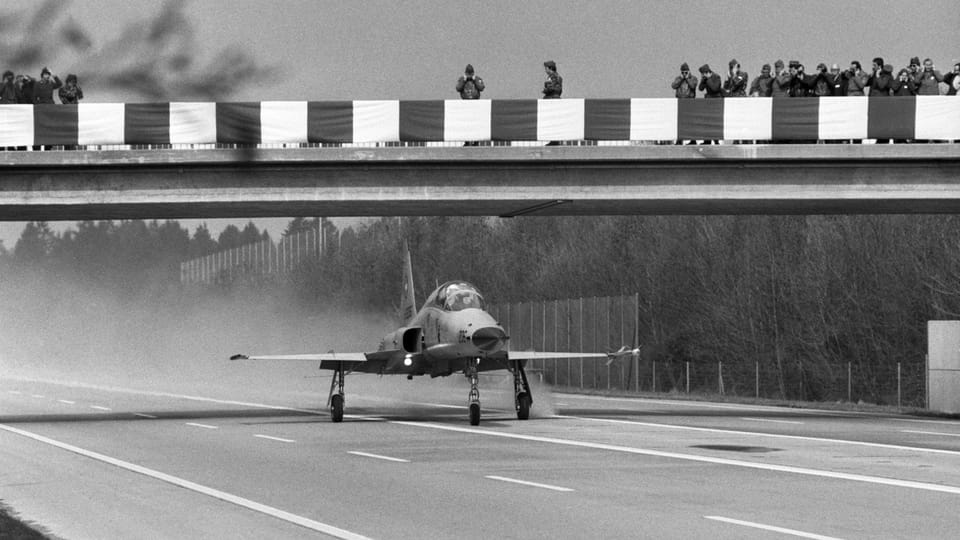 Kampfflugzeug landet auf Autobahn unter Beobachtung von Menschen auf einer Brücke.