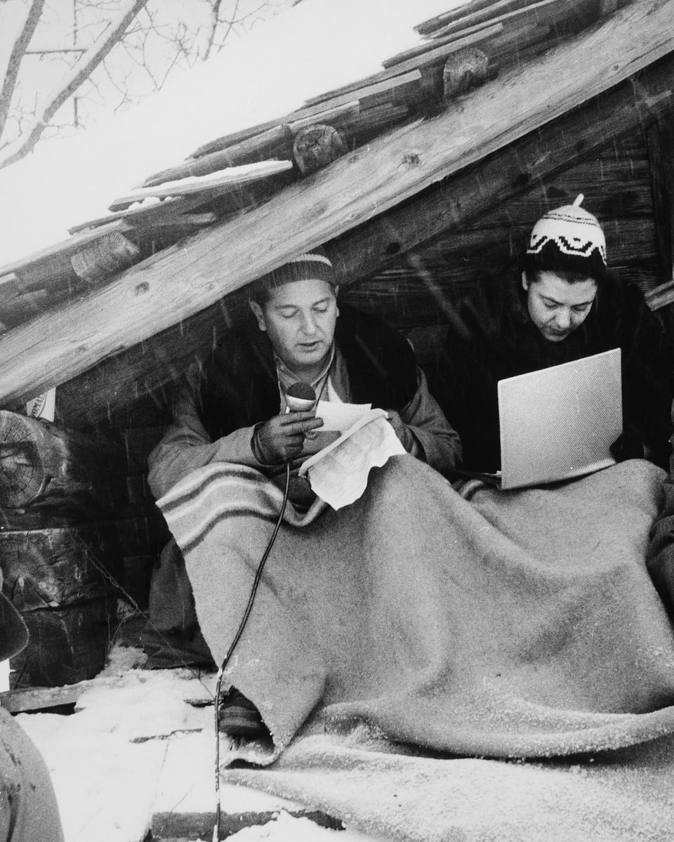 Die beiden Reporter sitzen unter einem Dachgiebel, schützen sich mit einer Wolldecke vor der Kälte und berichten live von den Rennen.
