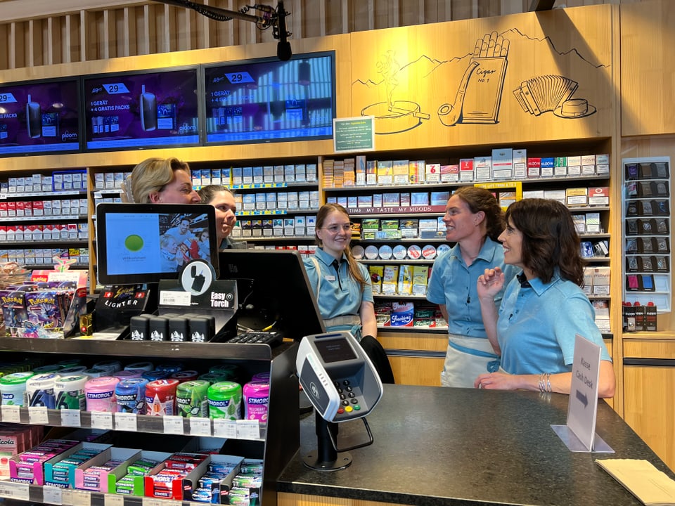 vier Frauen und Mona Vetsch, alle in der Uniform der Raststätte unterhalten sich hinter dem Tresen im Raststätten-Shop.