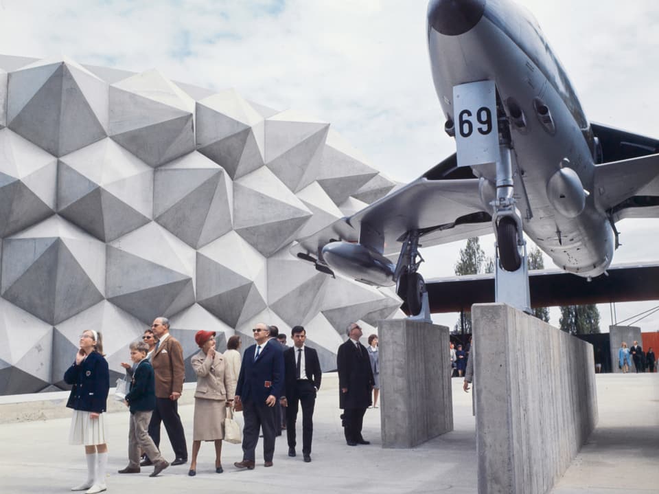 Menschen betrachten ein Flugzeug vor einem modernen Gebäude.