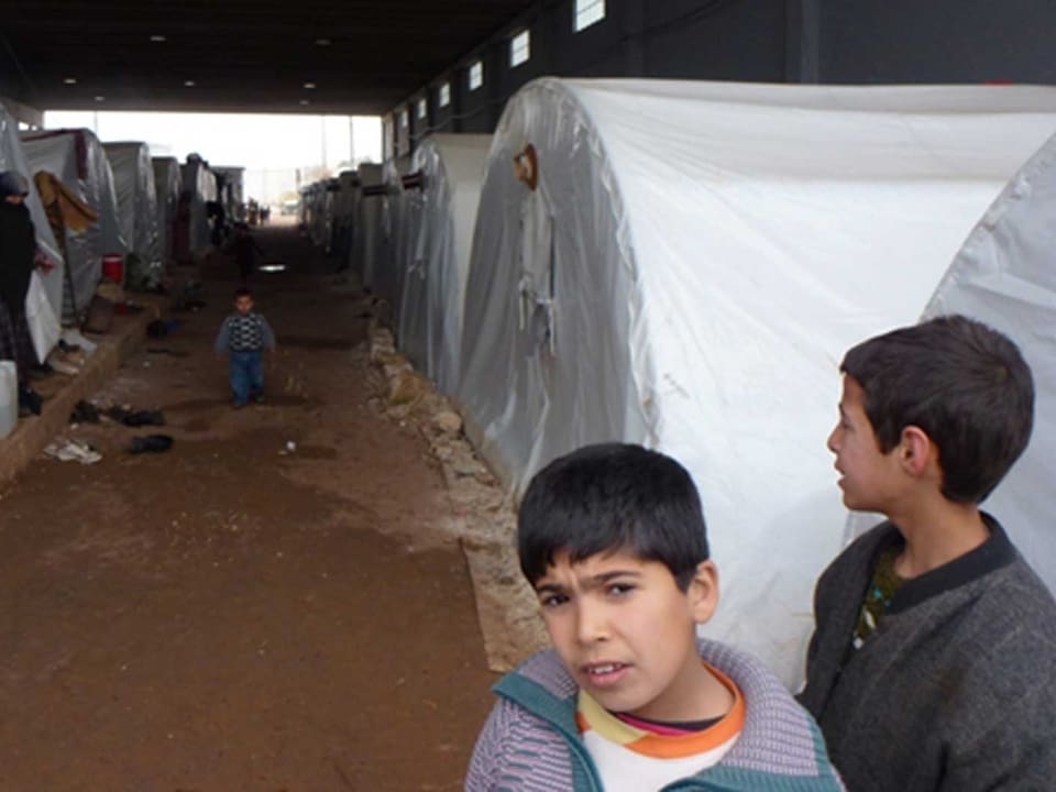 Zu sehen ist ein Flüchtlingslager. Eine Zeltgasse. Zwei Buben im Alter von ungefähr 7 Jahren gucken neugierig. Ein drittes Kind, ca. 3-jährig, läuft auf den Fotografen zu. Aus einem Zelt guckt eine Frau im Tschador.