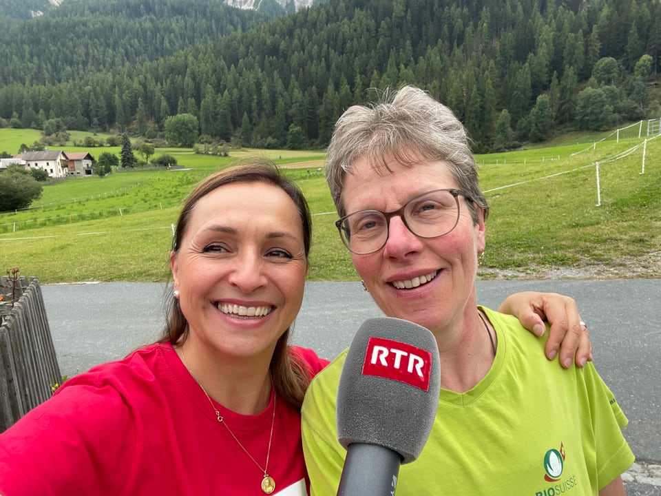 Zwei lächelnde Frauen, eine hält ein RTR-Mikrofon, Hintergrund mit grüner Landschaft und Wald.