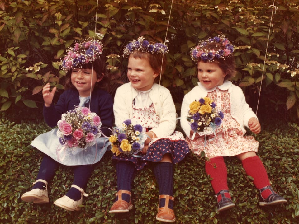 Drei kleine Mädchen sitzen auf einem Busch,tragen Blumenkränze auf dem Kopf und halten einen Blumenstrauss in der Hand.