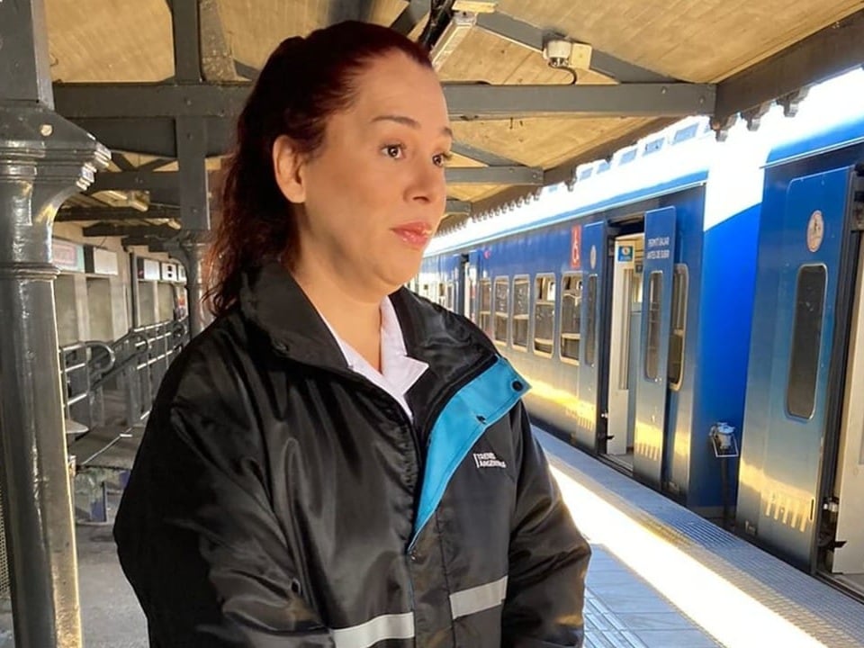 Frau in schwarzer Jacke steht an einem Bahngleis neben einem Zug.