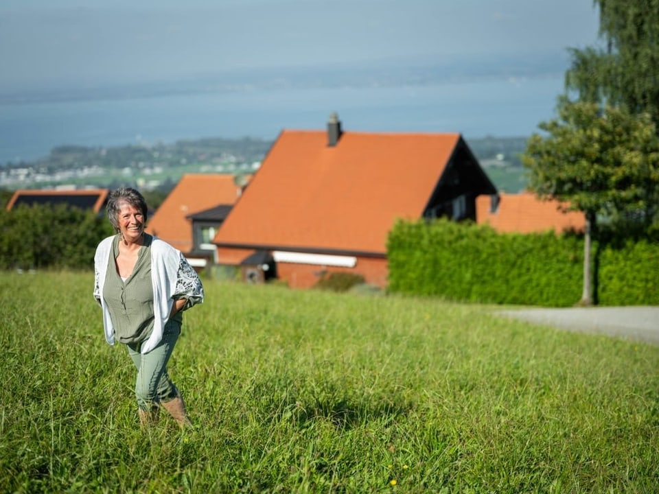 Frau auf Wiese vor Häusern mit rotem Dach und See im Hintergrund.