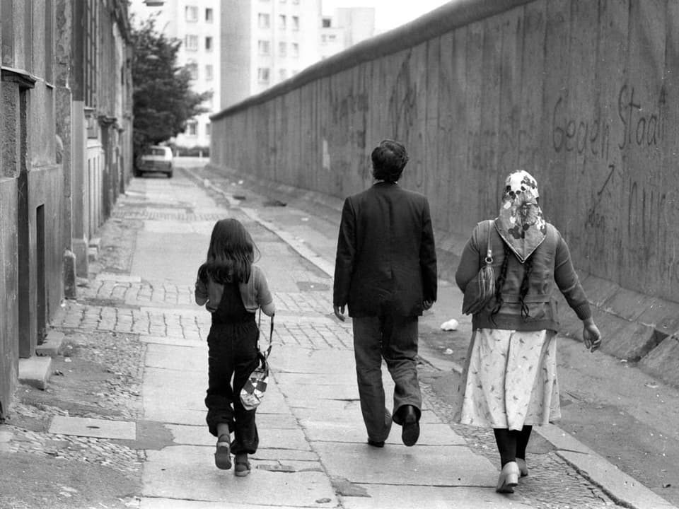 Schwarzweissaufnahme: Rückansicht einer türkischen Familie, die der Berliner Mauer entlangspaziert.