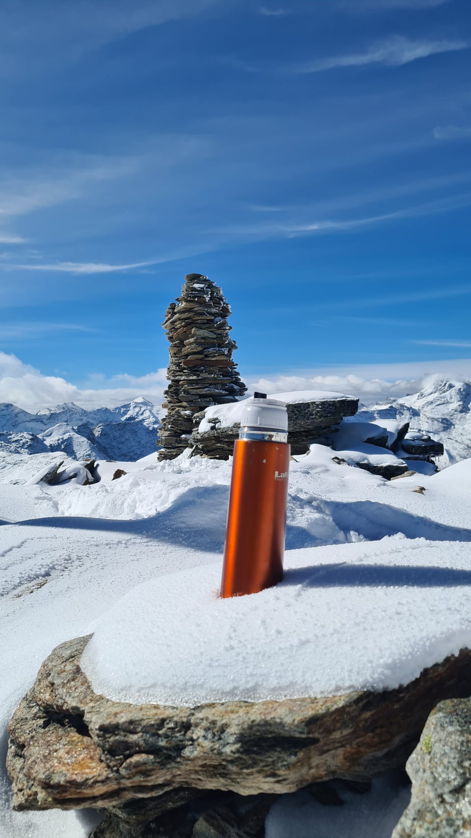 Thermoskanne auf verschneitem Berggipfel mit Steinmännchen im Hintergrund.