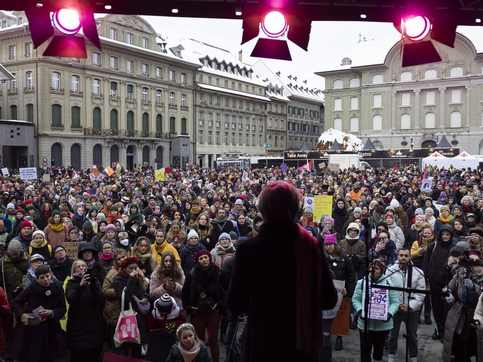 Sprecher auf Bühne vor grosser Menschenmenge bei Protest in Stadt.