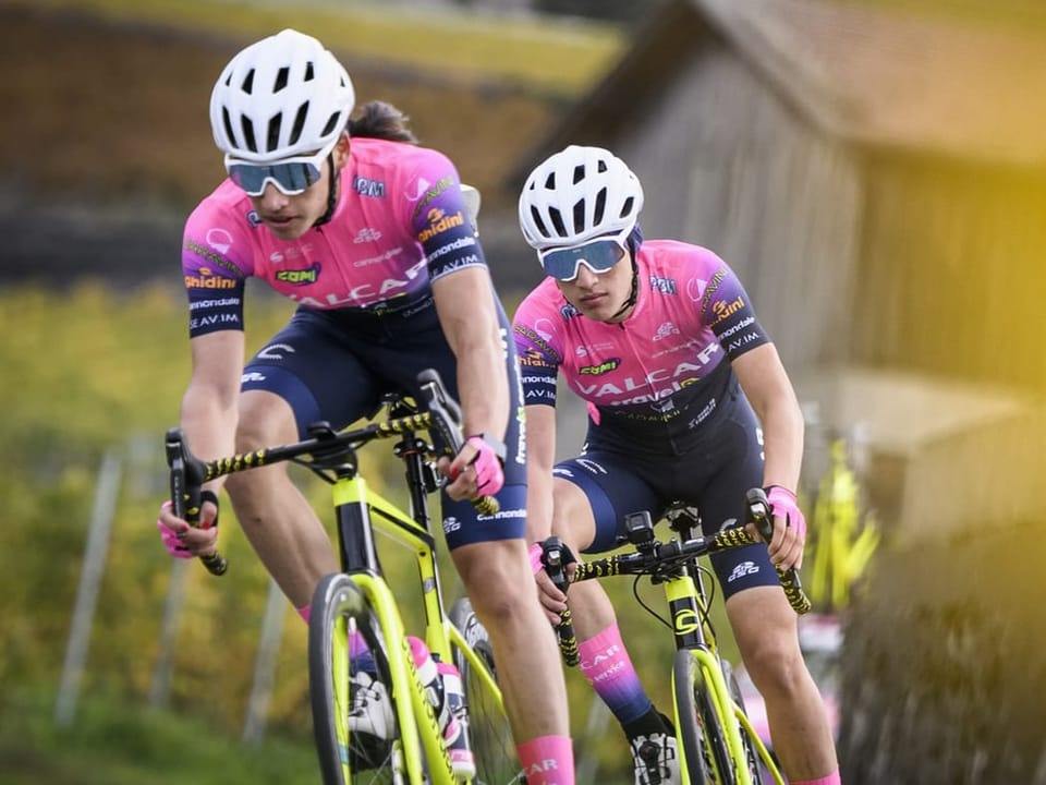Zwei Radfahrer in pink-blauen Trikots fahren draussen.