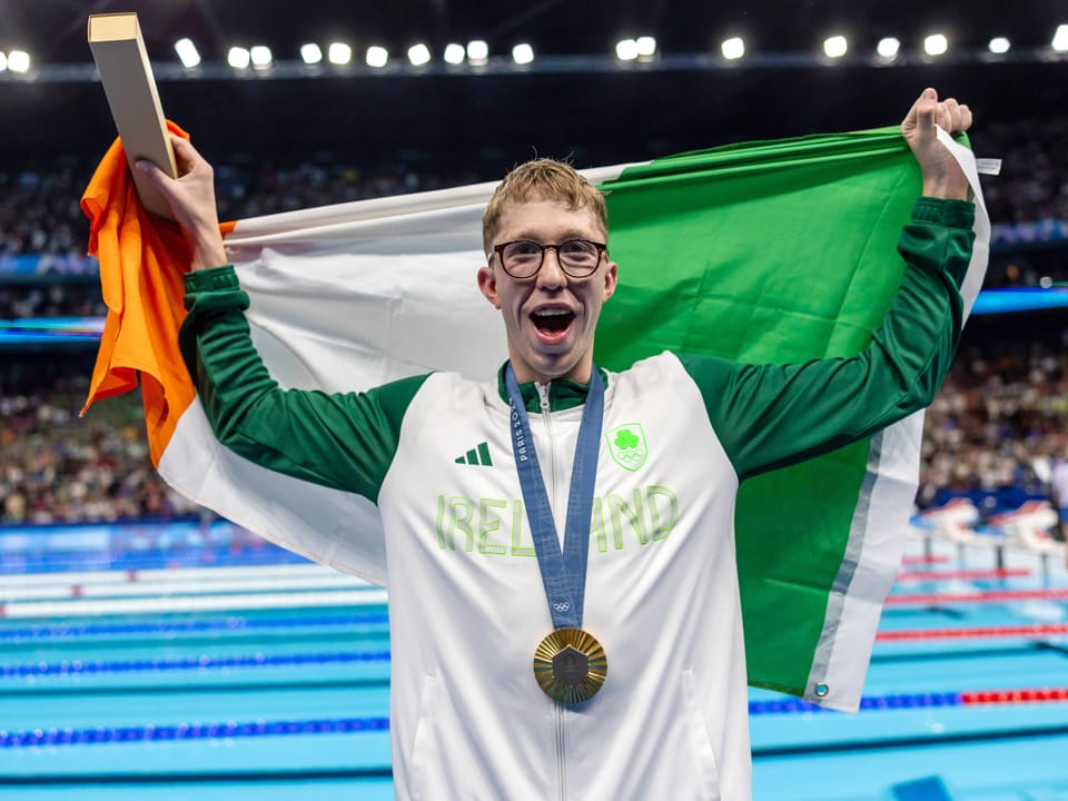 Schwimmer hält irische Flagge und trägt Medaille.