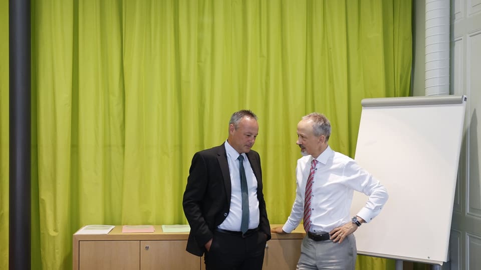 Zwei Männer im Gespräch vor grünem Vorhang und Flipchart.