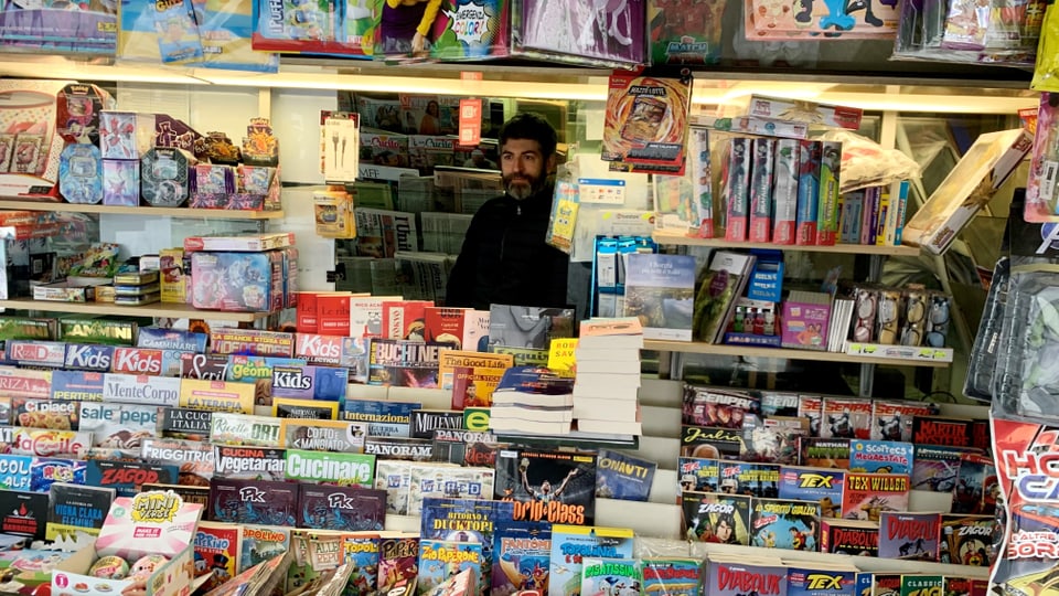 Mann steht hinter einem Kiosk voller Bücher und Zeitschriften.