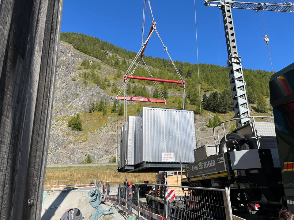 Ein grosser Container wird von einem Kran in einer bergigen Landschaft angehoben.