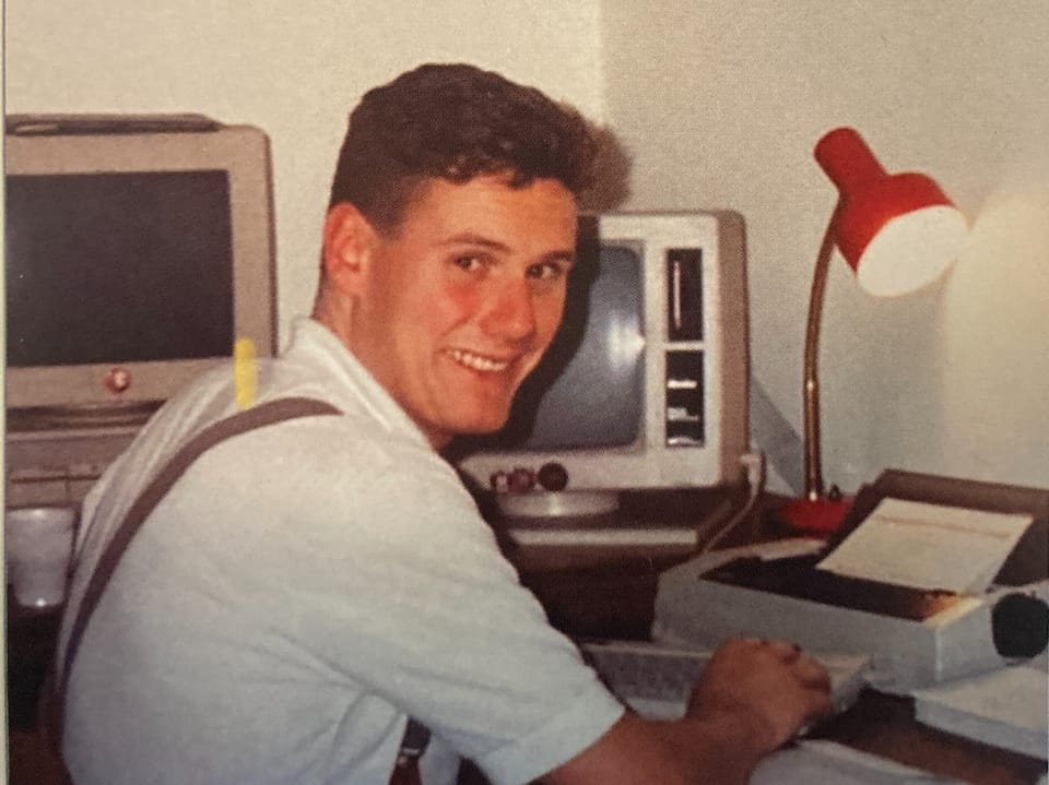 Lächelnder Mann an einem Schreibtisch mit einem alten Computer und einer Schreibmaschine.