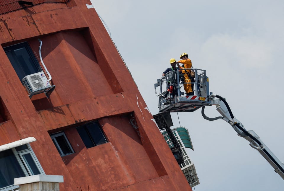 Rechts ein Feuerwehrs-Lift mit zwei Personen darauf, links dazu schräg stehendes Gebäude