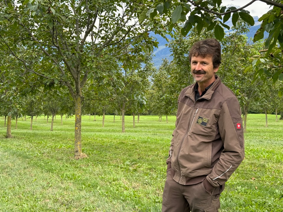 Mann in brauner Jacke steht in einem Obstgarten.