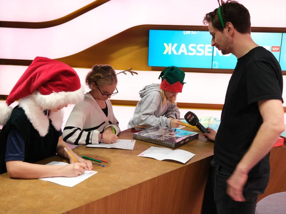 Drei Kinder schreiben etwas auf ein Blatt Papier auf. Moderator Raphi steht daneben mit einem Mikrofon. 