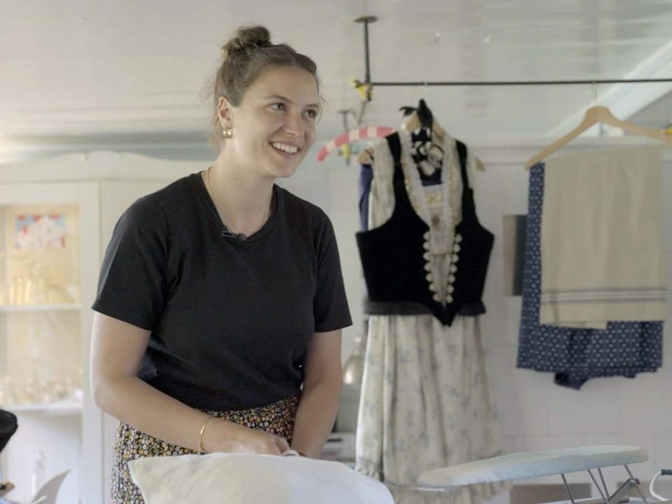 Junge Frau lächelt beim Bügeln in einem Zimmer mit traditioneller Kleidung im Hintergrund.