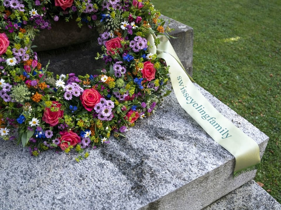 Blumenkranz auf einem Grabstein mit Schleife 'swisscyclingfamily'.